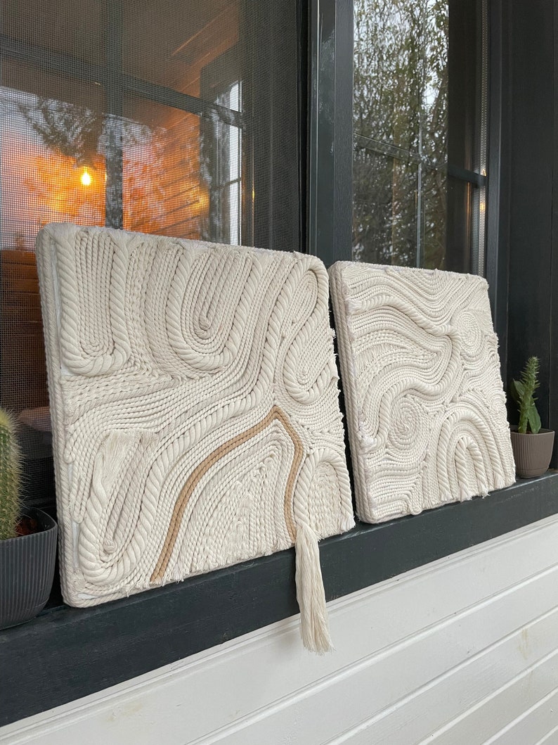 May include: Two white macrame wall hangings with a rope texture and a tassel on the bottom right hanging on a black and white wall.