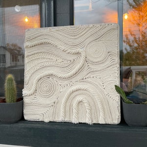 May include: A white wall hanging made of woven rope, creating a swirling pattern. The hanging is displayed on a windowsill with two potted cacti.