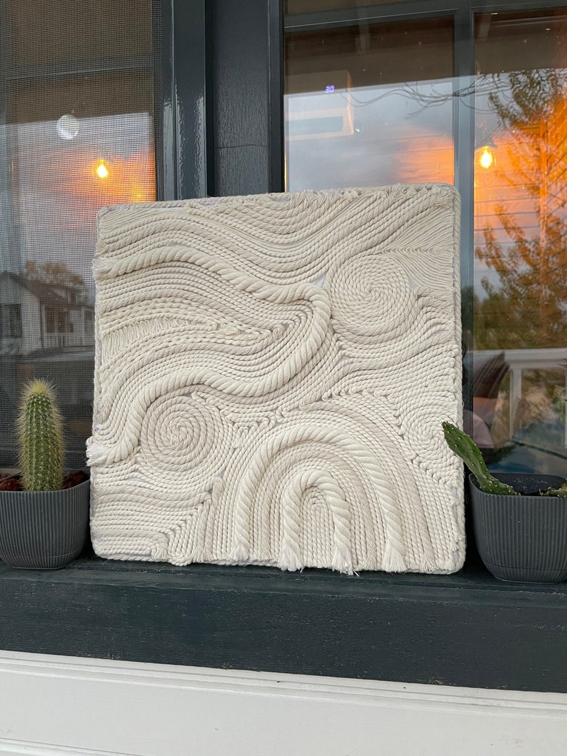 May include: A white wall hanging made of woven rope, creating a swirling pattern. The hanging is displayed on a windowsill with two potted cacti.