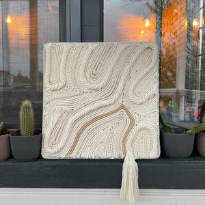 May include: A white macrame wall hanging with a brown rope design and a white tassel. The wall hanging is made of woven rope and has a textured surface.
