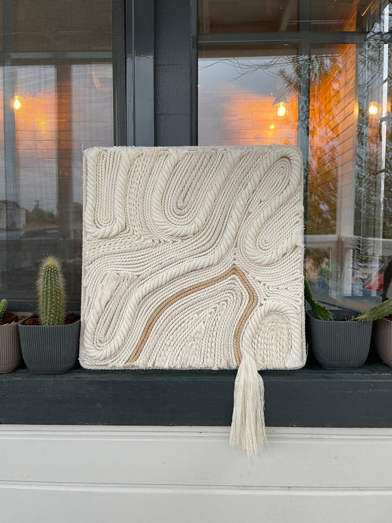 May include: A white macrame wall hanging with a brown rope design and a white tassel. The wall hanging is made of woven rope and has a textured surface.