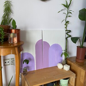 May include: Two wooden panels with a purple and white abstract design of arches. The panels are hanging on a white wall above a wooden shelf with potted plants.