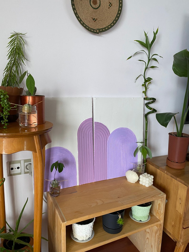 May include: Two wooden panels with a purple and white abstract design of arches. The panels are hanging on a white wall above a wooden shelf with potted plants.