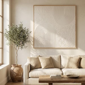 Op de afbeelding: Een vierkant kunstwerk aan de muur met een wit gestructureerd oppervlak met verhoogde boogpatronen, ingelijst in licht hout. Het kunstwerk toont een combinatie van gladde, concentrische halfronde lijnen en ruwe, onregelmatige secties.