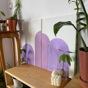 May include: Three purple painted arches on a white canvas background. The arches are textured and have a 3D effect. The canvas is leaning against a wooden shelf with plants and other decor.