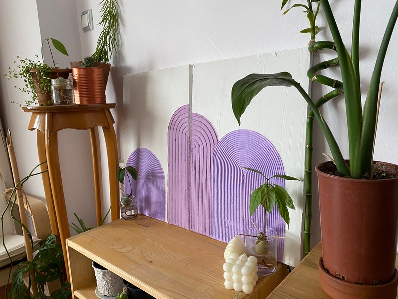 May include: Three purple painted arches on a white canvas background. The arches are textured and have a 3D effect. The canvas is leaning against a wooden shelf with plants and other decor.