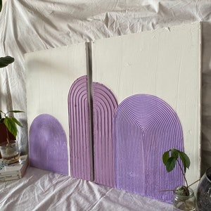 May include: Two white canvases with a textured surface, each featuring three purple, raised, semi-circular shapes. The shapes are arranged in a row, with the largest shape on the right and the smallest on the left. The shapes are made of a textured material that resembles a series of concentric circles.