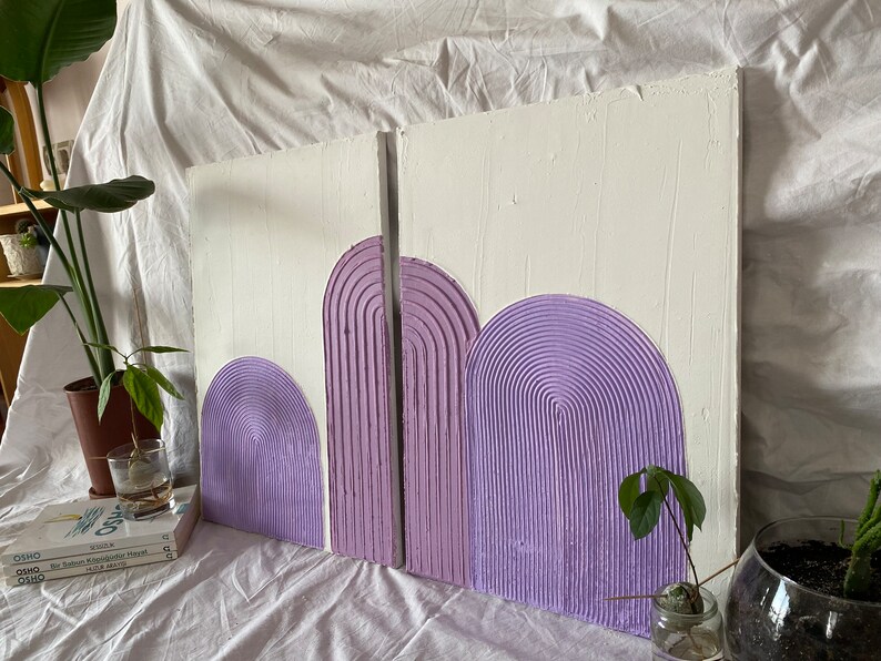 May include: Two white canvases with a textured surface, each featuring three purple, raised, semi-circular shapes. The shapes are arranged in a row, with the largest shape on the right and the smallest on the left. The shapes are made of a textured material that resembles a series of concentric circles.