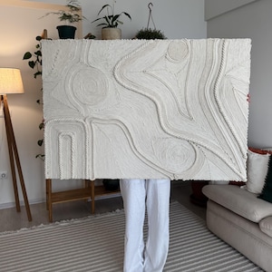 K&ouml;nnte beinhalten: Ein gro&szlig;er wei&szlig;er Wandbehang aus geflochtenem Seil, der ein strukturiertes abstraktes Design erzeugt. Der Behang wird von einer Person vor einer wei&szlig;en Wand gehalten.