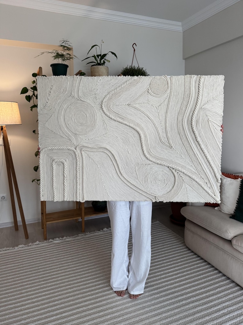 K&ouml;nnte beinhalten: Ein gro&szlig;er wei&szlig;er Wandbehang aus geflochtenem Seil, der ein strukturiertes abstraktes Design erzeugt. Der Behang wird von einer Person vor einer wei&szlig;en Wand gehalten.