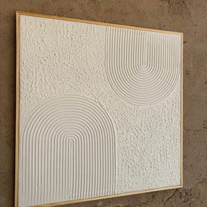 Op de afbeelding: Een vierkant wandkunstwerk met een wit gestructureerd oppervlak en twee verhoogde boogmotieven. De bogen bestaan uit parallelle verticale lijnen, terwijl de omliggende gebieden een ruwe, onregelmatige afwerking hebben. Het kunstwerk is gevat in een lichthouten lijst.