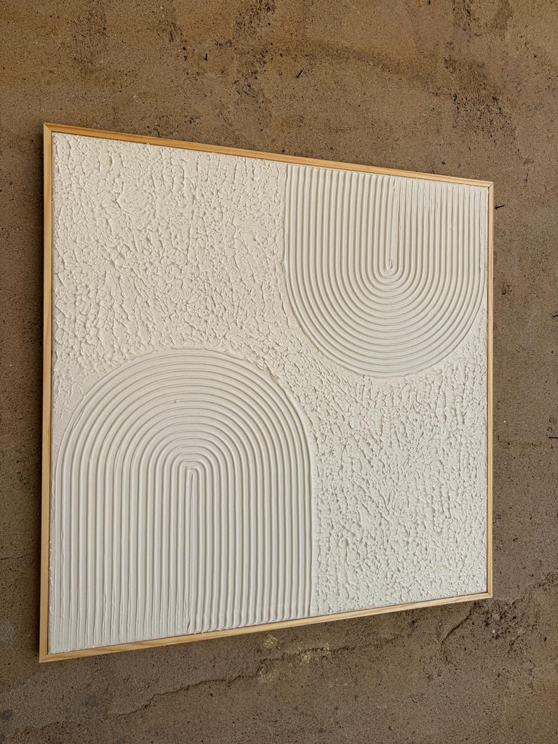 Op de afbeelding: Een vierkant wandkunstwerk met een wit gestructureerd oppervlak en twee verhoogde boogmotieven. De bogen bestaan uit parallelle verticale lijnen, terwijl de omliggende gebieden een ruwe, onregelmatige afwerking hebben. Het kunstwerk is gevat in een lichthouten lijst.