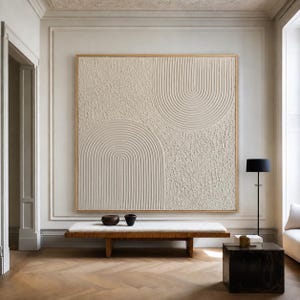 Op de afbeelding: Een groot vierkant wandkunstwerk met een getextureerd cr&egrave;mekleurig oppervlak met verhoogde, uitgesneden boogpatronen. Het kunstwerk is ingelijst in een dunne lichte houten rand en toont een combinatie van gladde groeven en ruwe, onregelmatige delen.