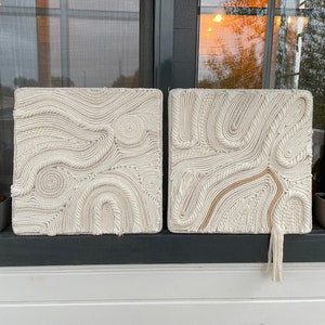 May include: Two white macrame wall hangings with a rope design. The wall hangings are square and have a fringe at the bottom.