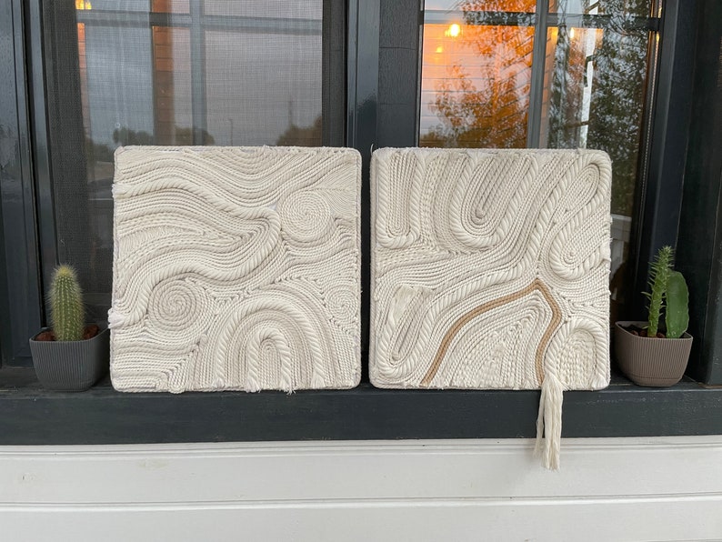 May include: Two white macrame wall hangings with a rope design. The wall hangings are square and have a fringe at the bottom.