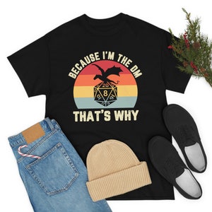 Op de afbeelding: Zwart T-shirt met een vintage-stijl grafische afbeelding van een draak en een 20-zijdige dobbelsteen. De tekst luidt "Because I'm the DM That's Why".