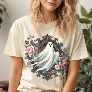 May include: A cream-colored t-shirt features a vintage-style illustration of a ghost amidst a floral arrangement. The ghost is depicted in soft, light blue and white hues, while the flowers are rendered in pastel pinks, purples, and greens. The design is framed by an ornate, dark-colored frame, adding a touch of elegance to the spooky theme. The t-shirt is a relaxed fit, suitable for casual wear.