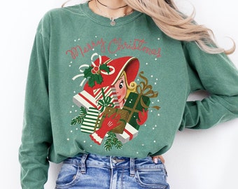 May include: A green long-sleeved sweatshirt with the text "Merry Christmas" and a vintage illustration of a woman in a red hat holding a stack of wrapped gifts.