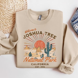 May include: A beige sweatshirt with a vintage-style graphic design featuring a sunset, mountains, and a cactus. The text reads "Welcome to Joshua Tree National Park, California, Est. 1994".