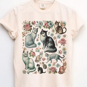 May include: A cream-colored short-sleeve t-shirt featuring a central graphic print of various cats in different poses interspersed with colorful floral illustrations including pink and blue flowers. The shirt has a crew neckline.
