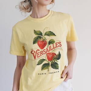 May include: A yellow t-shirt featuring a vintage-style graphic of strawberries and the word "Versailles" in red script. The design includes green leaves and the text "Paris, France". The shirt is paired with white shorts.