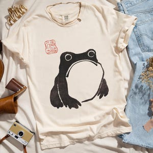 May include: A cream colored t-shirt with a black and white illustration of a frog. The frog is sitting on a white background with a red stamp that says "The Frog" in Japanese characters.