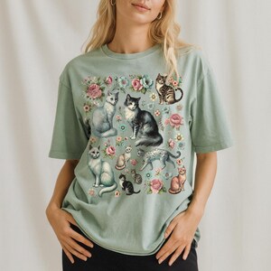 May include: A sage green short-sleeve t-shirt featuring a graphic print of various cats in different poses interspersed with colorful floral illustrations including pink and blue roses and small blossoms across the front chest area.