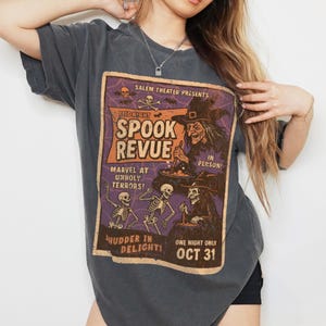 May include: Dark grey t-shirt with a vintage Halloween poster design. The poster reads "Midnight Spook Revue" with illustrations of skeletons and a witch. Text includes "Marvel at Unholy Terrors!" and "One Night Only Oct 31".