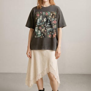 May include: A charcoal gray short-sleeve t-shirt featuring a graphic print of various cats surrounded by colorful flowers. The shirt is paired with an asymmetrical cream-colored skirt with a lace-trimmed hem and black lace-up boots.