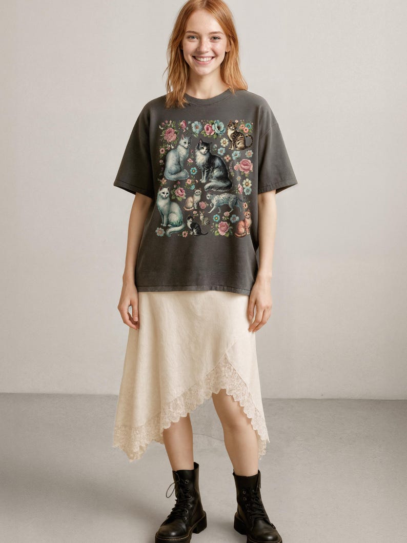 May include: A charcoal gray short-sleeve t-shirt featuring a graphic print of various cats surrounded by colorful flowers. The shirt is paired with an asymmetrical cream-colored skirt with a lace-trimmed hem and black lace-up boots.