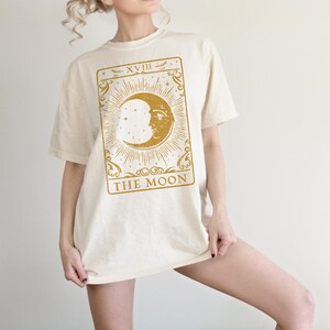 May include: A cream colored oversized t-shirt with a gold tarot card design featuring the moon and the text "XVIII THE MOON".