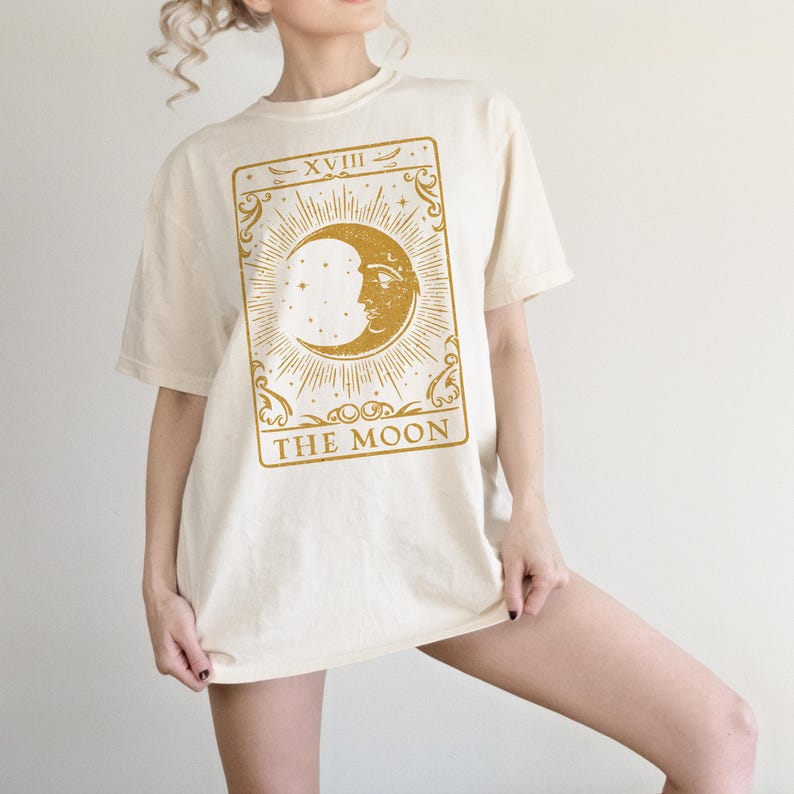 May include: A cream colored oversized t-shirt with a gold tarot card design featuring the moon and the text "XVIII THE MOON".