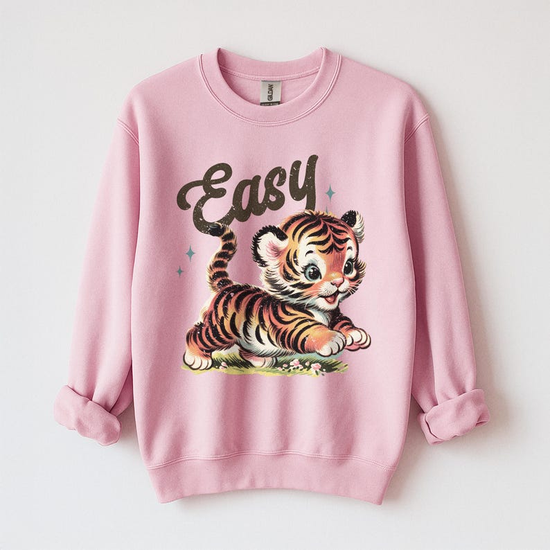 Puede incluir: Una sudadera rosa claro con una ilustraci&oacute;n de estilo vintage de un lindo cachorro de tigre con rayas negras y naranjas. La palabra "Easy" est&aacute; escrita encima del tigre en una fuente marr&oacute;n y cursiva. La sudadera tiene cuello redondo y mangas largas.