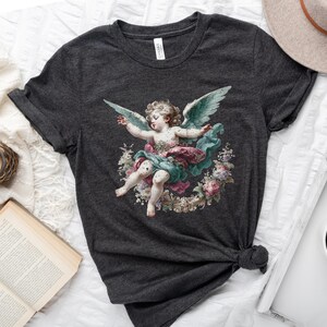 Puede incluir: Una camiseta gris oscuro con una ilustraci&oacute;n vintage de un querub&iacute;n con alas, rodeado de flores. El querub&iacute;n lleva un vestido rosa y verde.