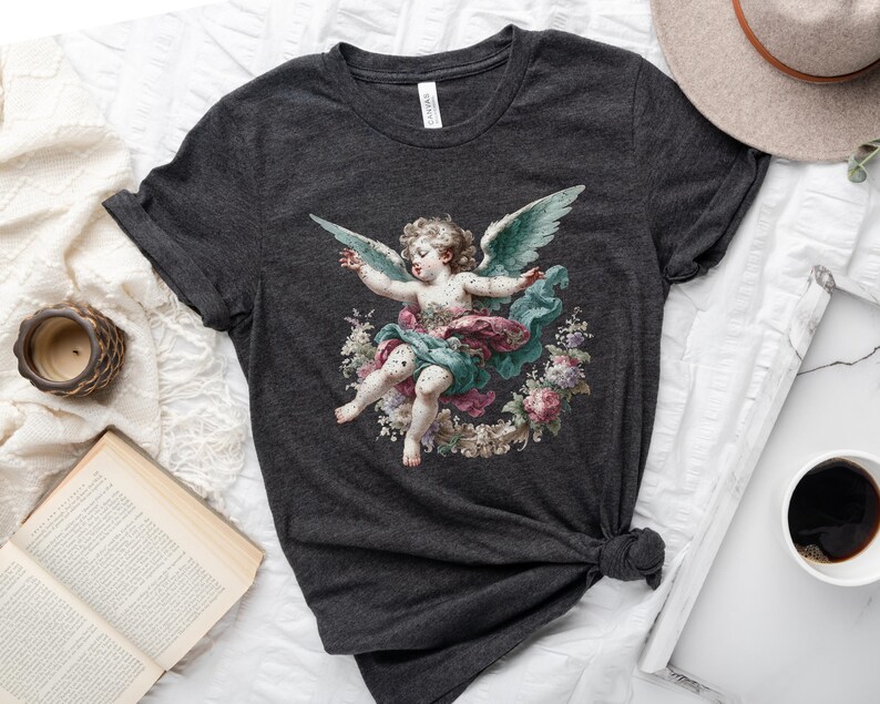 Puede incluir: Una camiseta gris oscuro con una ilustraci&oacute;n vintage de un querub&iacute;n con alas, rodeado de flores. El querub&iacute;n lleva un vestido rosa y verde.