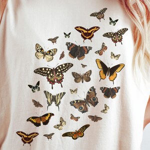 K&ouml;nnte beinhalten: Ein wei&szlig;es T-Shirt mit einem Muster aus bunten Schmetterlingen. Die Schmetterlinge sind in verschiedenen Gr&ouml;&szlig;en und Formen und in einem zuf&auml;lligen Muster angeordnet.