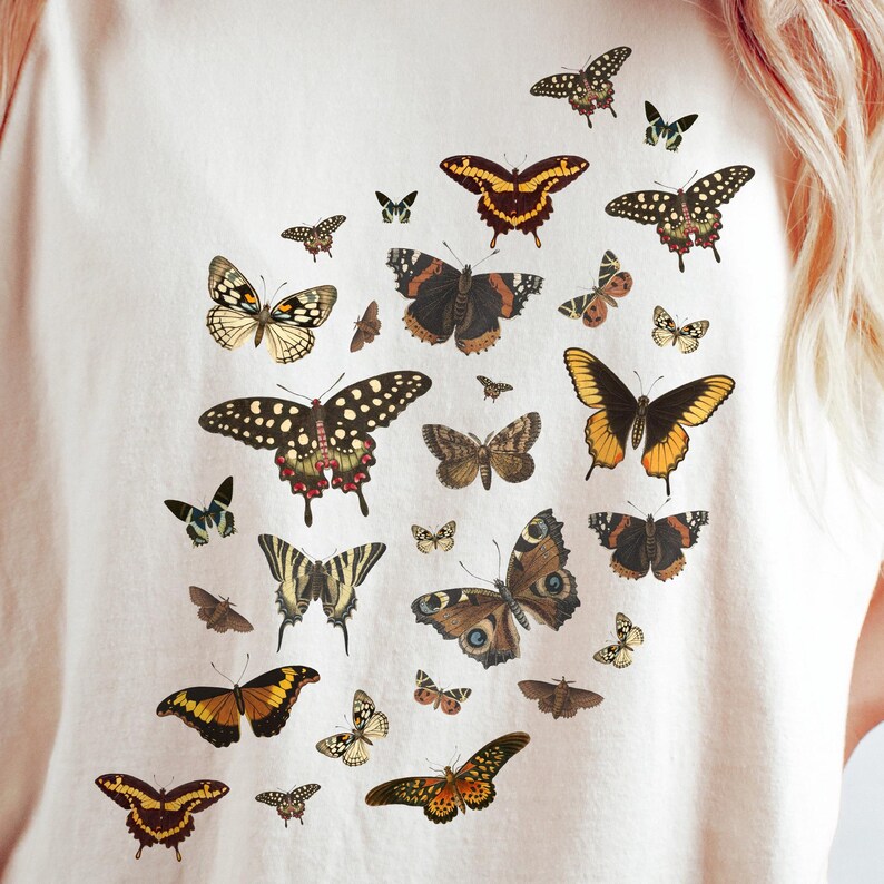 K&ouml;nnte beinhalten: Ein wei&szlig;es T-Shirt mit einem Muster aus bunten Schmetterlingen. Die Schmetterlinge sind in verschiedenen Gr&ouml;&szlig;en und Formen und in einem zuf&auml;lligen Muster angeordnet.