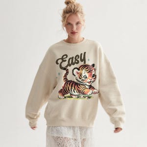 Puede incluir: Sudadera de color crema con una ilustraci&oacute;n de un tigre de dibujos animados con rayas negras y naranjas. La palabra "Easy" est&aacute; escrita encima del tigre. La sudadera tiene cuello redondo y mangas largas.