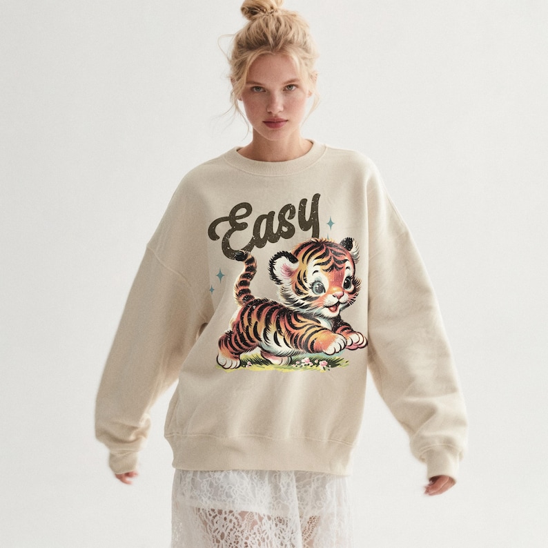 Puede incluir: Sudadera de color crema con una ilustraci&oacute;n de un tigre de dibujos animados con rayas negras y naranjas. La palabra "Easy" est&aacute; escrita encima del tigre. La sudadera tiene cuello redondo y mangas largas.