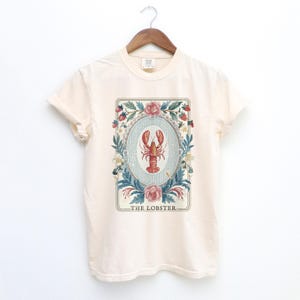 K&ouml;nnte beinhalten: Cremefarbenes T-Shirt mit einer detaillierten Illustration eines roten Hummers in einem ovalen Rahmen. Der Rahmen ist von floralen Elementen umgeben, darunter Rosen, Erdbeeren und Bl&auml;tter. Der Text "THE LOBSTER" ist darunter gedruckt.