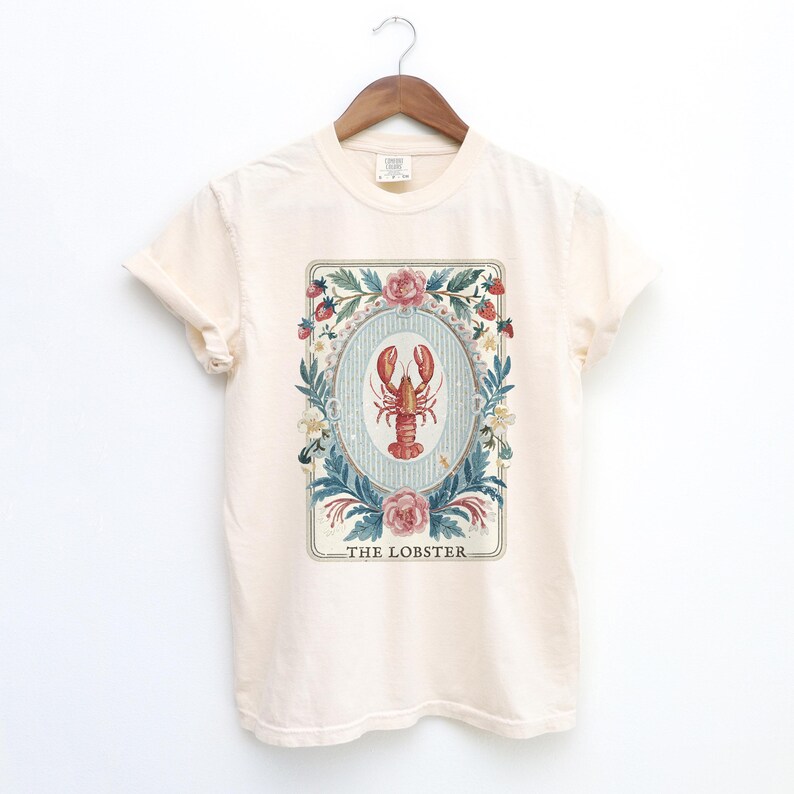 K&ouml;nnte beinhalten: Cremefarbenes T-Shirt mit einer detaillierten Illustration eines roten Hummers in einem ovalen Rahmen. Der Rahmen ist von floralen Elementen umgeben, darunter Rosen, Erdbeeren und Bl&auml;tter. Der Text "THE LOBSTER" ist darunter gedruckt.