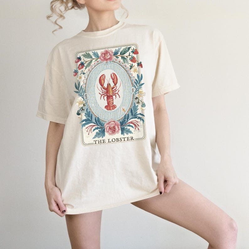 K&ouml;nnte beinhalten: Cremefarbenes T-Shirt mit einer gerahmten Illustration eines roten Hummers in einem blauen Oval, umgeben von Blumen und Erdbeeren. Der Text "THE LOBSTER" befindet sich unten. Das Shirt hat eine lockere Passform.