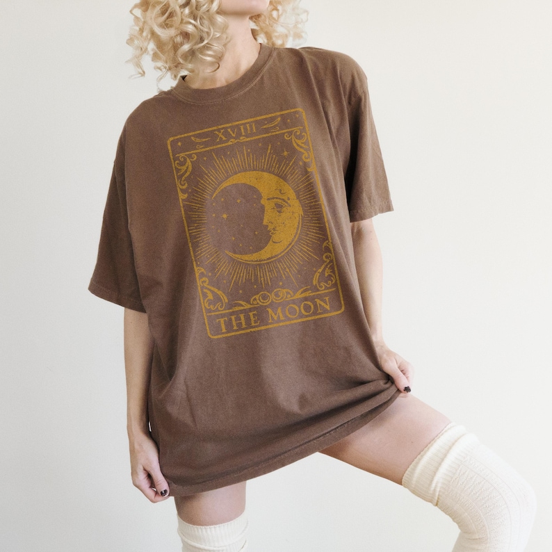 May include: A brown oversized t-shirt with a vintage tarot card design featuring the moon and the text "XVIII THE MOON".
