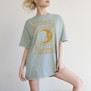 May include: A light blue oversized t-shirt with a gold tarot card design featuring the moon and the text "XVIII THE MOON".