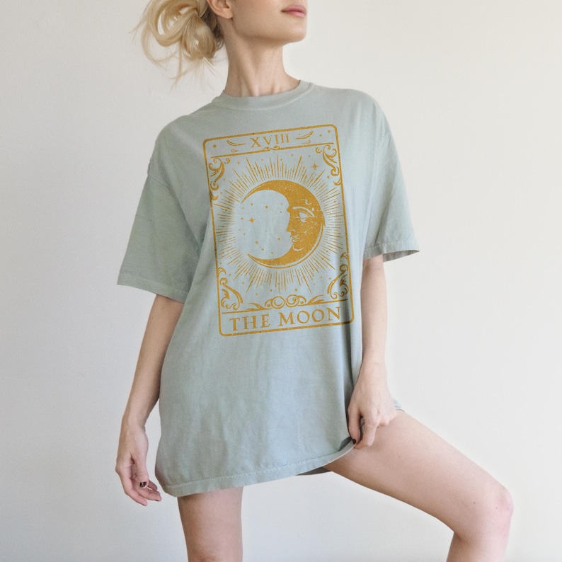 May include: A light blue oversized t-shirt with a gold tarot card design featuring the moon and the text "XVIII THE MOON".
