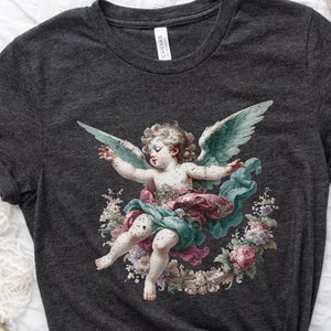 Puede incluir: Una camiseta gris oscuro con un estampado vintage de un querub&iacute;n con alas, rodeado de flores. El querub&iacute;n est&aacute; pintado en tonos de azul, rosa y verde.