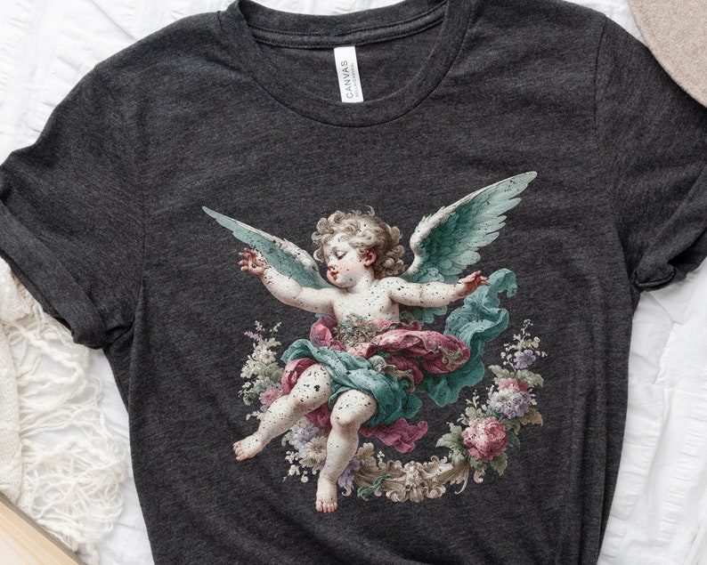 Puede incluir: Una camiseta gris oscuro con un estampado vintage de un querub&iacute;n con alas, rodeado de flores. El querub&iacute;n est&aacute; pintado en tonos de azul, rosa y verde.