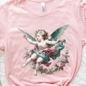 Puede incluir: Una camiseta rosa con una ilustraci&oacute;n vintage de un querub&iacute;n con alas, rodeado de flores. El querub&iacute;n lleva un vestido blanco y una corona de flores en la cabeza.