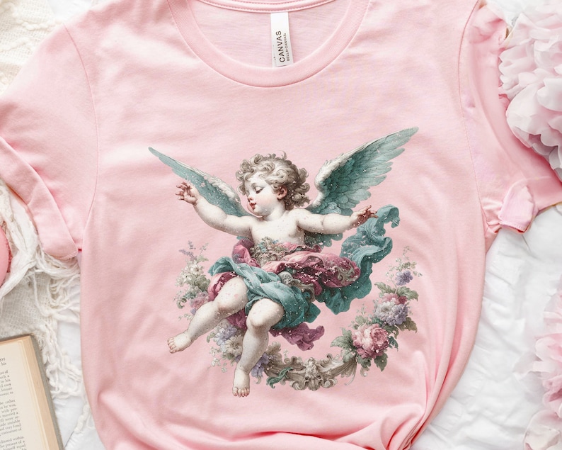 Puede incluir: Una camiseta rosa con una ilustraci&oacute;n vintage de un querub&iacute;n con alas, rodeado de flores. El querub&iacute;n lleva un vestido blanco y una corona de flores en la cabeza.