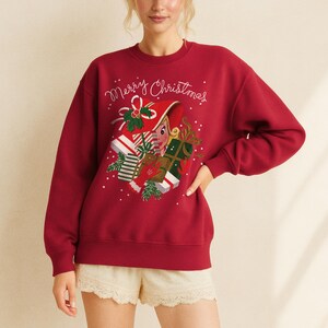 May include: A burgundy sweatshirt with the embroidered text "Merry Christmas." The design features a festive elf with wrapped gifts. The sweatshirt is a solid color and has a crew neck.